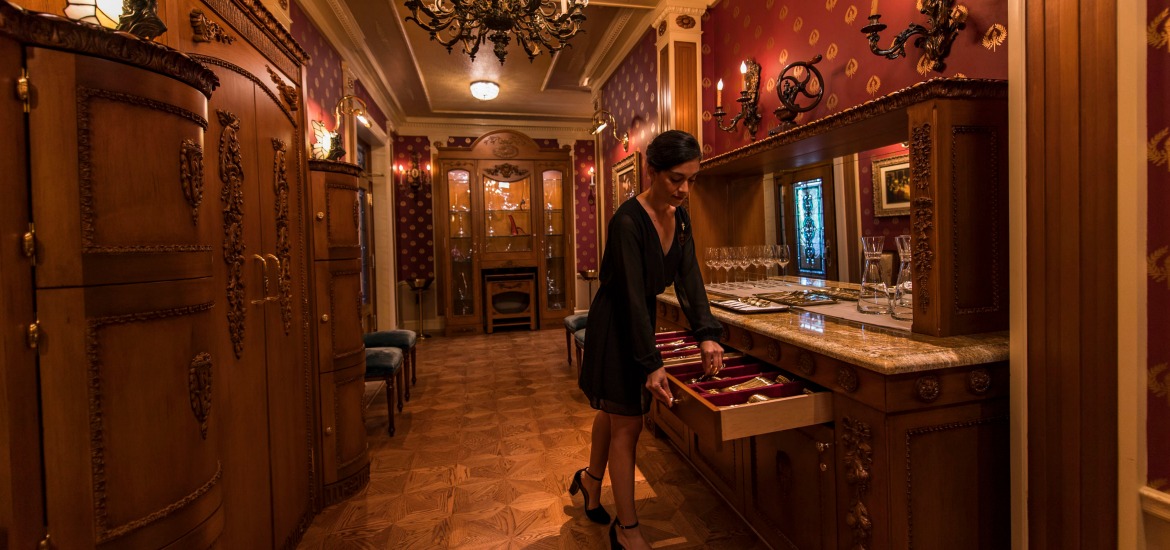 A female hostess prepares silverware for a dinner service at a mirrored silverware sideboard in a room with wooden cabinets and glass displays